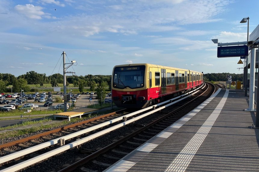 Das Foto zeigt einen Bahnsteig mit S-Bahn sowie im Hintergrund parkende Autos.