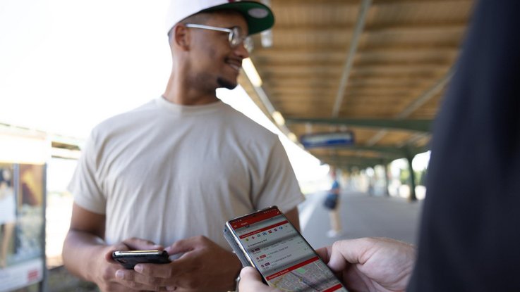 Zwei Personen am Bahnsteig. Eine davon hält ein Smartphone mit der geöffneten App.